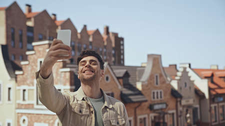 cheerful man taking selfie with blurred buildings on background