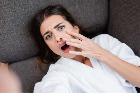 top view of scared young adult woman looking at camera with open mouth and hand near face on gray couchの写真素材