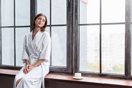 pretty young adult woman in bathrobe sitting on sill with cup of coffee near window at homeの写真素材