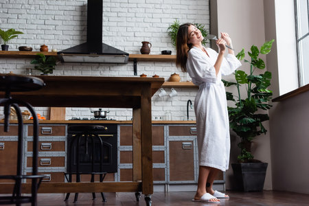 playful young adult woman in white bathrobe holding spatula and singing in modern loftの写真素材