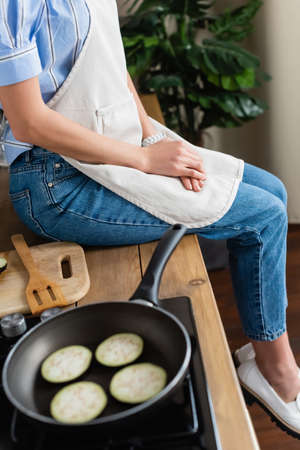 partial view of young adult woman in apron sitting near spot with slices of eggplant in modern kitchenの写真素材