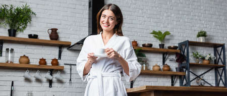 pretty young adult woman in bathrobe drinking coffee and looking at camera in modern loft, bannerの写真素材