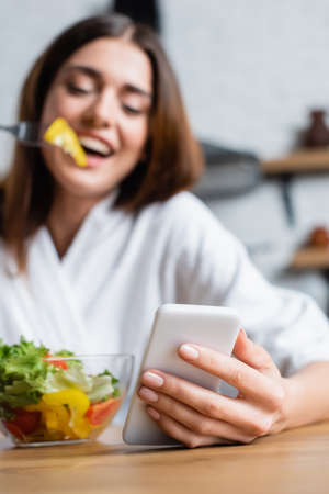 cheerful young adult woman in bathrobe eating salad and using cellphone in modern kitchen, focus on foregroundの写真素材