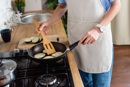 cropped view of young adult woman frying slices of eggplant in pan and using spatula in kitchenの写真素材