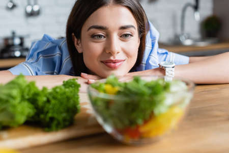 smiling young adult woman looking at prepared vegetables salad in kitchenの写真素材