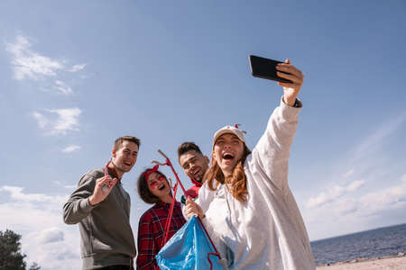 cheerful woman taking selfie with friends while picking up trashの写真素材