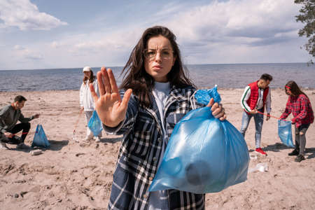 young woman showing stop gesture near blurred group of volunteers picking up trash, ecology conceptの写真素材