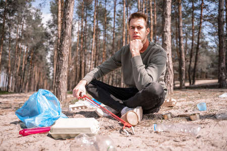 pensive man with crossed legs sitting near garbage on ground in woodsの写真素材