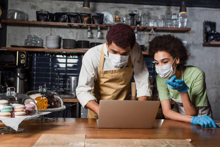 African american barista in medical mask pointing at laptop near colleague in cafeの写真素材