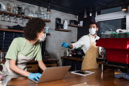 African american barista in medical mask standing near blurred colleague using laptop in cafeの写真素材