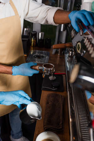 Cropped view of african american barista in latex gloves holding coffee in holder near coffee machine and colleagueの写真素材