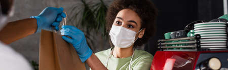 African american barista giving paper bag to colleague in latex gloves in cafe, bannerの写真素材