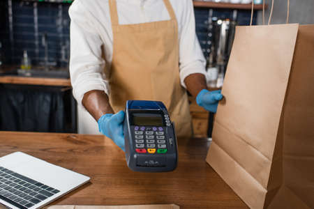 Cropped view of african american barista in latex gloves holding payment terminal and paper bag in cafeの写真素材
