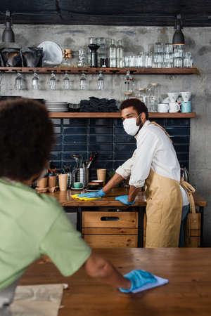 African american barista in medical mask and latex gloves cleaning worktop with rag near blurred...