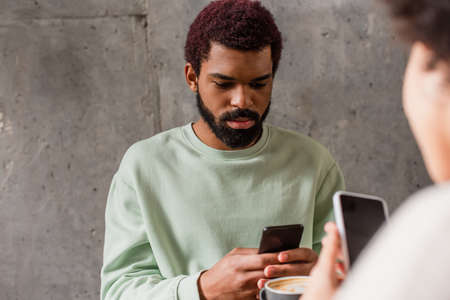 African american man using cellphone near cup of coffee and blurred girlfriend in cafeの写真素材