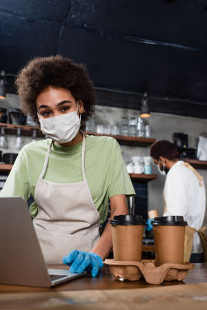 African american barista in medical mask and latex glove using laptop near coffee in cafeの写真素材