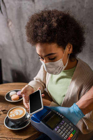 African american client in medical mask paying with smartphone near waiter with terminal in cafe
