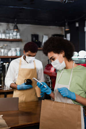 African american barista in medical mask using smartphone and holding paper bag near blurred...