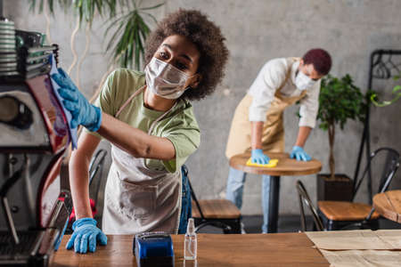 African american barista in medical mask cleaning coffee machine near payment terminal and...