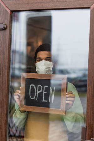 African american barista in medical mask holding chalkboard with open lettering near door of cafeの写真素材