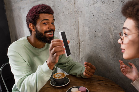Positive african american man holding smartphone near blurred girlfriend and coffee in cafe