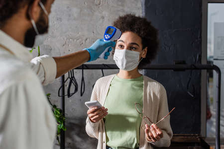 African american client in medical mask holding smartphone near waiter with infrared thermometer on...