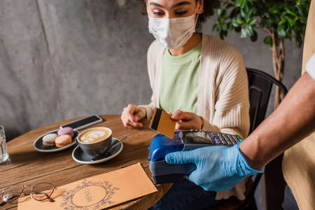 African american waiter holding payment terminal near client with credit card on blurred background