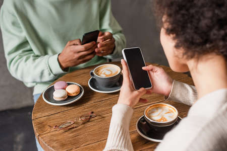 Smartphone in hands of african american woman near cappuccino and blurred boyfriend in cafe