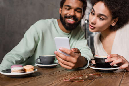 Smartphone in hand of smiling african american man near girlfriend with coffee and macaroons in cafeの写真素材