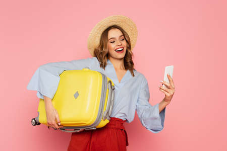Smiling woman with suitcase using smartphone isolated on pinkの写真素材