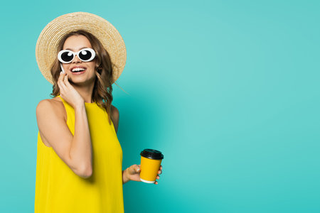 Cheerful woman in sunglasses holding coffee in paper cup and talking on cellphone on blue backgroundの写真素材