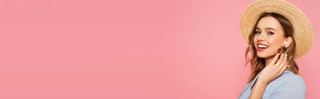 Cheerful woman in sun hat and earphone looking at camera isolated on pink, bannerの写真素材