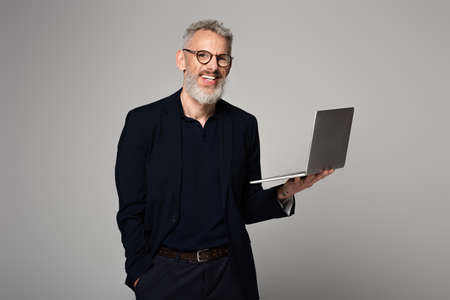 happy man with gray hair holding laptop while standing with hand in pocket isolated on gray