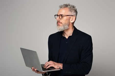 pensive middle aged man with gray hair in suit holding laptop isolated on grayの写真素材