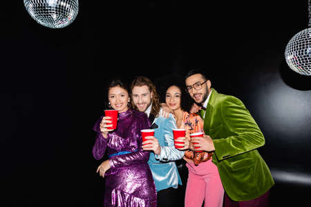 funny multiracial friends in colorful clothes standing with plastic cups in night club on black backgroundの写真素材
