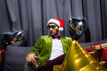 young adult arab man sitting on couch in santa hat and holding glass of champagne on gray backgroundの写真素材