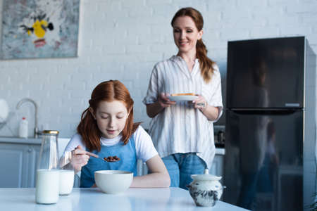 happy woman holding sandwich near preteen daughter eating tasty corn flakes for breakfast