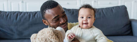 cheerful baby looking at camera near african american father and teddy bear, bannerの写真素材