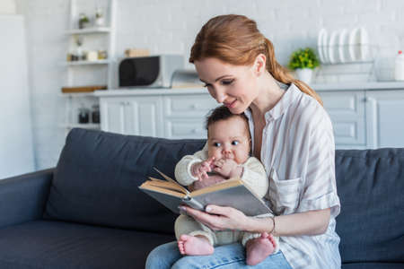 woman reading book to african american baby girl while sitting on couch in kitchen