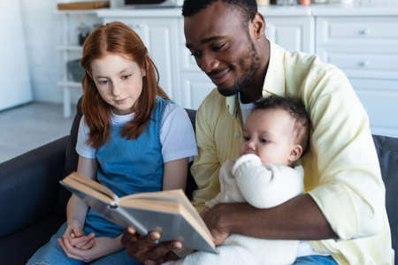 smiling african american man reading book to interracial daughters at homeの写真素材