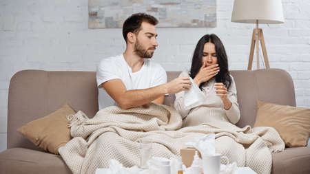 caring man giving tissue to sick woman sneezing in living roomの写真素材