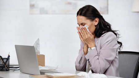 allergic woman sneezing in tissue near laptop on desk in officeの写真素材
