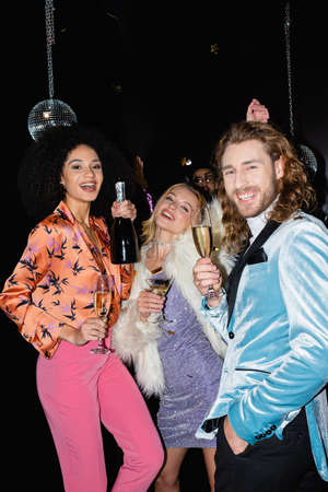 positive multiracial friends in colorful clothes dancing with champagne in night club on black backgroundの写真素材