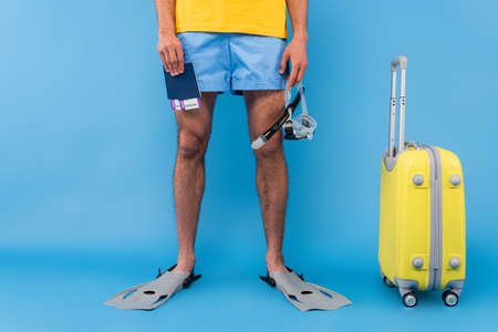 Cropped view of man in swimming flippers holding passports and goggles near suitcase on blue backgroundの写真素材