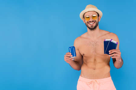 Cheerful shirtless man in sunglasses holding canned drink and passports isolated on blueの写真素材