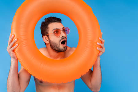 Excited man in sunglasses holding inflatable ring isolated on blueの写真素材