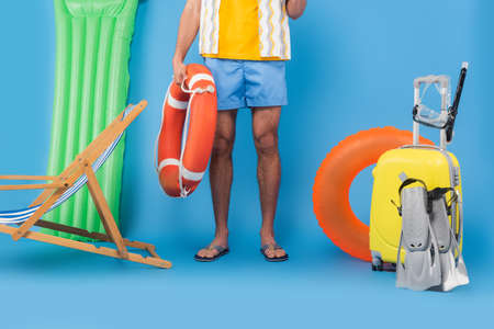 Cropped view of man holding life buoy near deck chair, suitcase and swimming flippers on blue...