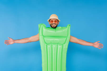 Smiling man in sun hat and sunglasses standing near inflatable mattress isolated on blueの写真素材