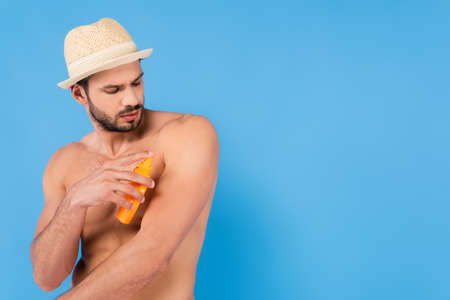 Muscular man in sun hat applying sunscreen isolated on blueの写真素材