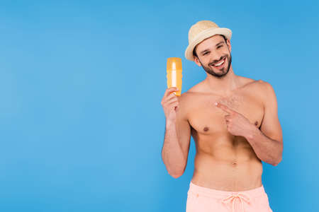 Shirtless man in sun hat pointing at sunscreen isolated on blueの写真素材
